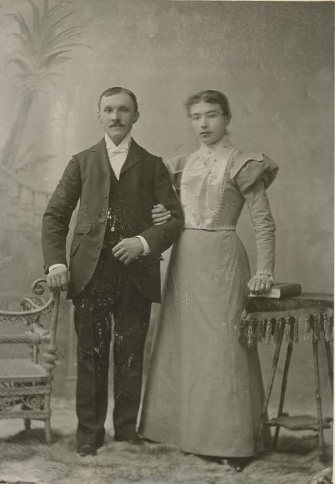 chair, cabinet photo, Iowa History, history of Iowa, Iowa, Portraits - Group, Olsson, Ann and Jons, couple, book, Cedar Falls, IA, painted backdrop, watch chain, tablecloth, wicker chair, table